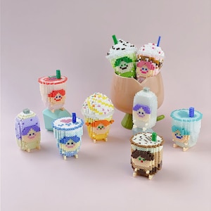 Może przedstawiać: Kolekcja kolorowych, pikselowanych rzeźb napojów, w tym wzory bubble tea i mrożonej kawy. Rzeźby mają różne kolory i wzory, ze szczegółami takimi jak słomki i bita śmietana. Są ułożone na różowym tle.
