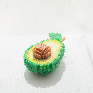 May include: A green avocado made of plastic beads, with a brown pit and a green stem. The avocado is half-cut, showing the pit inside.