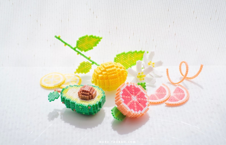 May include: A still life of beaded fruit, including a yellow lemon, a green avocado, and a pink grapefruit. The fruit is arranged on a white surface with a lemon branch and white flowers.
