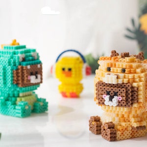 May include: Three small, colorful, pixelated figurines made of beads. The figurines are a green dinosaur, a yellow chick, and a brown bear wearing a yellow and orange costume.