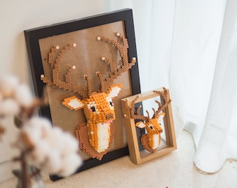 May include: Two framed wall decorations featuring brown and white beaded deer heads. The larger deer head is in a black frame and the smaller deer head is in a wooden frame.