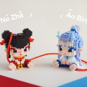 Puede incluir: Dos pequeñas figuras de personajes de dibujos animados hechas de cuentas de colores. La figura de la izquierda es negra con detalles rojos y está etiquetada como "Ne Zha". La figura de la derecha es azul con detalles blancos y está etiquetada como "Ao Bing".