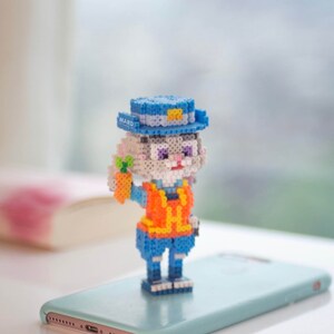 May include: A small, white, rabbit-like figure wearing a blue hat with a yellow badge that says "MARD" and an orange vest. The figure is holding a small orange carrot in its right hand. The figure is standing on a light blue smartphone.