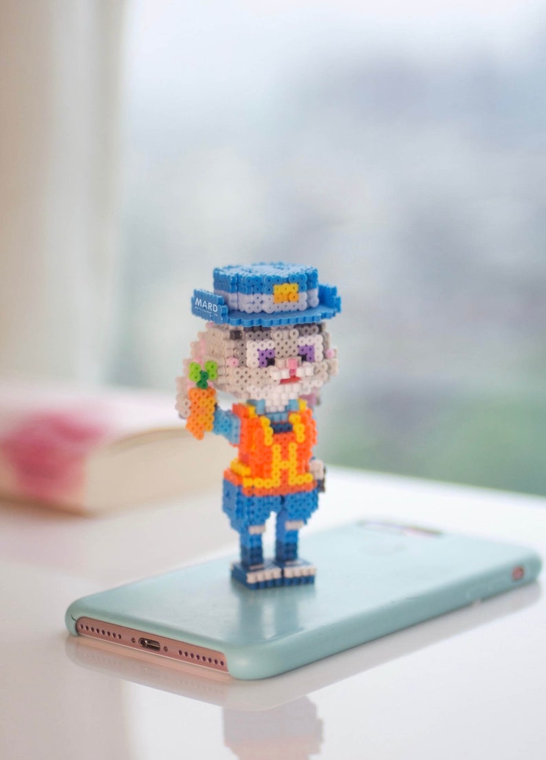 May include: A small, white, rabbit-like figure wearing a blue hat with a yellow badge that says "MARD" and an orange vest. The figure is holding a small orange carrot in its right hand. The figure is standing on a light blue smartphone.