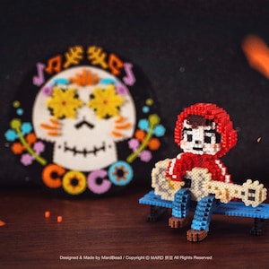 Può includere: Una figura pixelata di un personaggio del film Coco, seduto su una panchina blu che suona la chitarra. Sullo sfondo c'è un teschio colorato con la scritta "COCO". La figura indossa una felpa con cappuccio rossa e jeans blu.