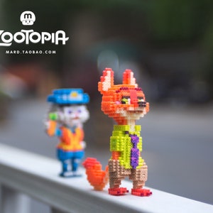 May include: Two small, colorful, pixelated figurines of characters from the movie Zootopia. The figurine on the left is a bunny wearing a police uniform and holding a carrot. The figurine on the right is a fox wearing a green shirt and a purple tie. The figurines are standing on a white railing.
