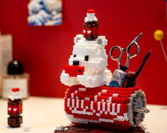 May include: A white polar bear figurine holding a red and white Coca-Cola can. The bear is wearing a red Santa hat and has a brown bottle on top of its head. The can is filled with various items, including a pair of scissors, a tube of hand cream, and a mascara wand.