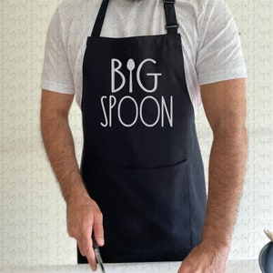 Puede incluir: Un delantal negro con un diseño blanco que dice "BIG SPOON" con un gráfico de cuchara sobre la palabra "BIG".