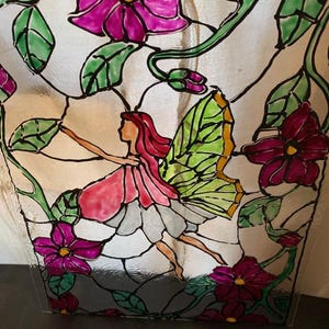 May include: A stained glass panel featuring a central fairy figure with red hair and green wings, surrounded by vibrant magenta flowers and green vines. The design uses black lead lines to define the shapes of the petals, leaves, and fairy.