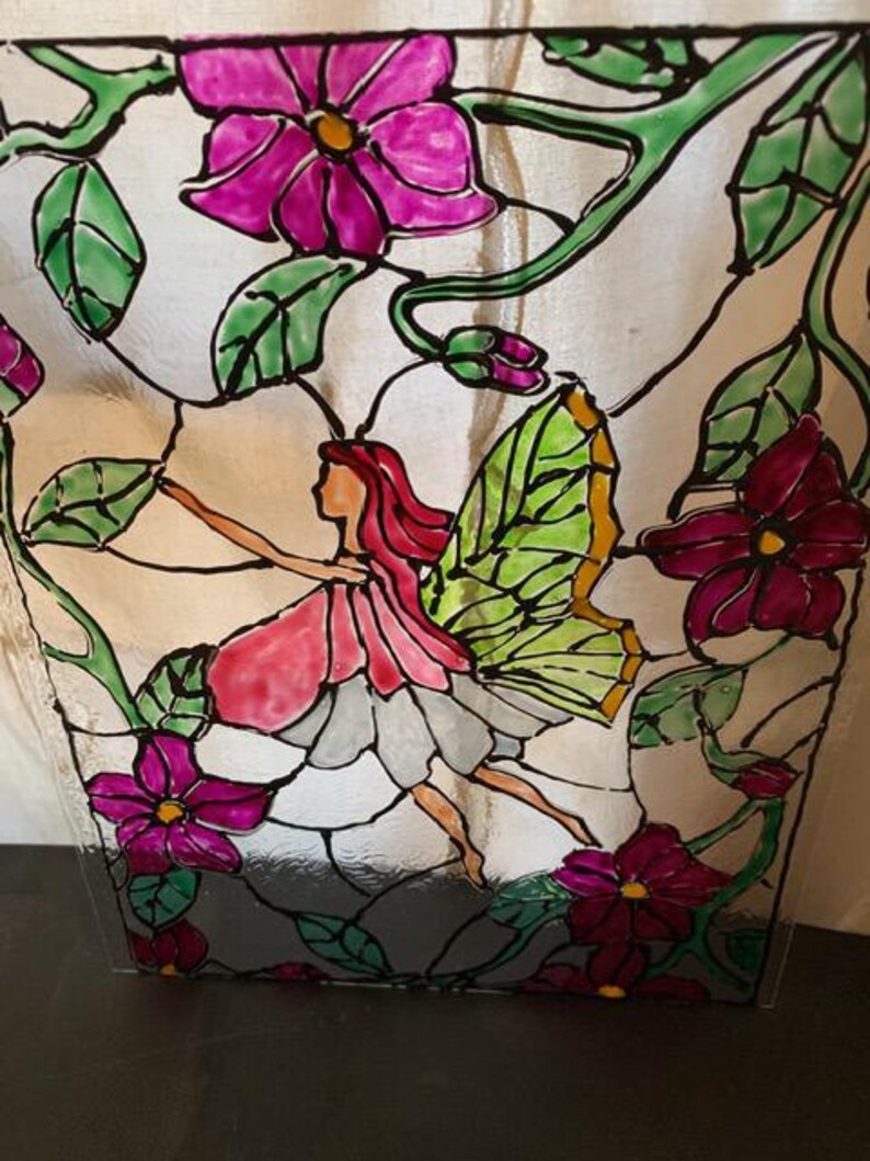 Pu&ograve; includere: Un pannello in vetro colorato raffigurante una figura di fata centrale con capelli rossi e ali verdi, circondata da vivaci fiori magenta e viti verdi. Il design utilizza linee di piombo nere per definire le forme dei petali, delle foglie e della fata.