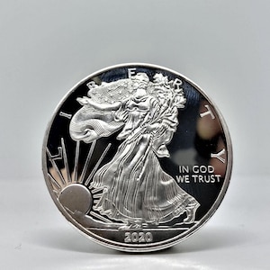 Può includere: Una moneta d'argento American Eagle con l'anno 2020 inciso sul fondo. La moneta presenta una raffigurazione di Lady Liberty che cammina con una torcia e un ramo d'ulivo. L'iscrizione "In God We Trust" è visibile sul lato destro della moneta.