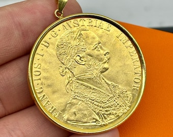 Könnte beinhalten: Ein goldfarbener Anhänger mit einem detaillierten Profil einer historischen Figur. Der Anhänger hat eine runde Form mit geprägtem Text am Rand. Die Öse ist ebenfalls goldfarben.
