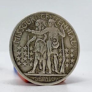 May include: Two silver coins, one with a portrait of a woman and the text "United States of America" and "Half Dollar", the other with the text "Missouri Centennial" and "Sedalia" and a depiction of three figures.