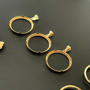 K&ouml;nnte beinhalten: Drei goldfarbene Metall-M&uuml;nzenanh&auml;nger-Bezel mit einer runden &Ouml;ffnung. Die Bezel haben eine glatte, polierte Oberfl&auml;che und sind so konzipiert, dass sie eine M&uuml;nze oder ein anderes rundes Objekt aufnehmen k&ouml;nnen.