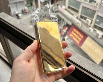 Puede incluir: Un objeto rectangular de color dorado envuelto en plástico transparente. El objeto está siendo sostenido en una mano.