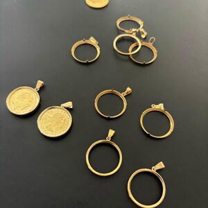 K&ouml;nnte beinhalten: Eine Sammlung von goldfarbenen M&uuml;nzanh&auml;ngern und Rahmen. Die Anh&auml;nger sind rund und haben oben eine &Ouml;se. Die Rahmen sind ebenfalls rund und haben einen Rand um den Rand. Die Rahmen sind daf&uuml;r ausgelegt, M&uuml;nzen zu halten.