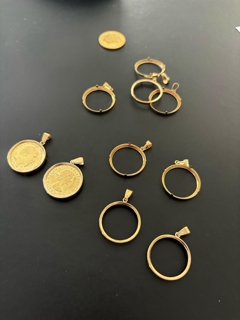 K&ouml;nnte beinhalten: Eine Sammlung von goldfarbenen M&uuml;nzanh&auml;ngern und Rahmen. Die Anh&auml;nger sind rund und haben oben eine &Ouml;se. Die Rahmen sind ebenfalls rund und haben einen Rand um den Rand. Die Rahmen sind daf&uuml;r ausgelegt, M&uuml;nzen zu halten.