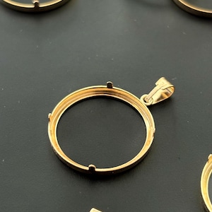 Puede incluir: Pendiente de bisel de moneda de metal dorado con un bucle para sujetar una cadena. El bisel está diseñado para sujetar una moneda o un medallón redondo.