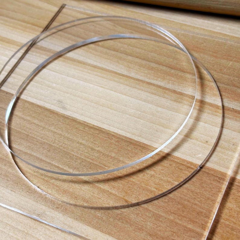 May include: Clear, thin, circular plastic pieces, possibly for crafting or jewelry making, are arranged on a light brown wooden surface. The plastic appears transparent, with a slight sheen, and is curved into circles.