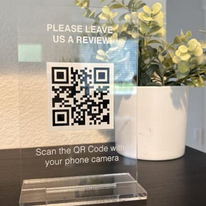 May include: A clear acrylic sign featuring the white text PLEASE LEAVE US A REVIEW at the top and Scan the QR Code with your phone camera at the bottom, with a central black and white QR code square mounted on a rectangular acrylic base.