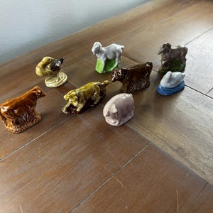 May include: A collection of ceramic farm animal figurines, including a cow, pig, sheep, duck, and dog. The figurines are painted in various colors, such as brown, white, yellow, and pink. The figurines are arranged on a wooden surface.