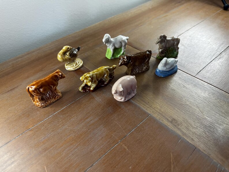 May include: A collection of ceramic farm animal figurines, including a cow, pig, sheep, duck, and dog. The figurines are painted in various colors, such as brown, white, yellow, and pink. The figurines are arranged on a wooden surface.