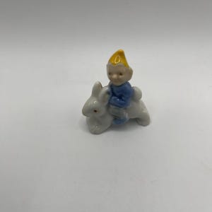 May include: A small, white ceramic figurine of a gnome riding a rabbit. The gnome wears a yellow hat and blue shirt. The rabbit has its ears perked up and is in a running position. The figurine is on a white background.