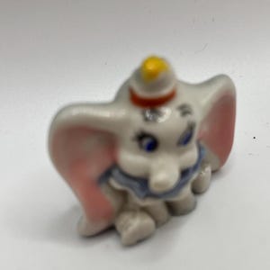 Puede incluir: Una pequeña figurita de cerámica de Dumbo el elefante de Dumbo de Disney. La figurita es blanca con orejas rosas y un collar azul y blanco. Dumbo lleva un sombrero amarillo y rojo.