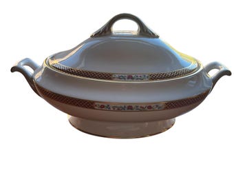 May include: A white ceramic serving dish with a lid and handles. The dish features a decorative band of red and gold geometric patterns, with floral accents. The lid has a gold handle.
