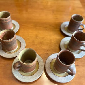 May include: Six handmade ceramic coffee cups and saucers. The cups are brown with a textured finish and small handles. The saucers are beige with a yellow centre. The set is arranged on a wooden surface.