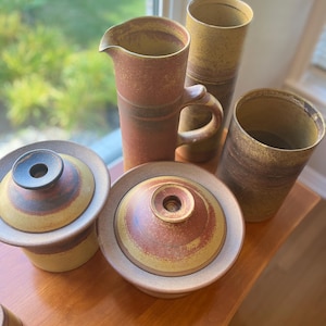 Puede incluir: Colección de cerámica artesanal en tonos marrones, amarillos y rojos. El conjunto incluye una jarra con asa, dos recipientes con tapa y dos jarrones cilíndricos. La cerámica tiene una estética rústica y terrosa.