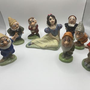Può includere: Una collezione di statuette in ceramica raffiguranti i personaggi della fiaba di Biancaneve. Il set comprende Biancaneve, sette nani e altri personaggi. Le statuette sono dipinte con colori vivaci e sono esposte su una superficie bianca.
