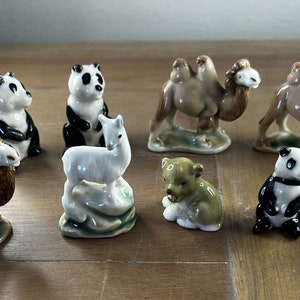 Puede incluir: Una colección de nueve figuritas de animales en miniatura de cerámica, que incluyen un panda, un camello, una llama, un oso y un pájaro. Las figuritas están pintadas en colores realistas y se exhiben sobre una superficie de madera.