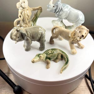 Puede incluir: Un conjunto de cinco figuritas de animales de cerámica, que incluyen un mono, un elefante, un rinoceronte, un león y un caimán. Las figuritas están pintadas en colores realistas y están en una variedad de poses.