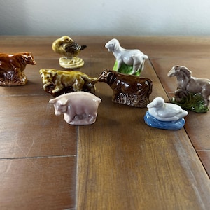 May include: A collection of miniature ceramic farm animal figurines, including a cow, duck, sheep, pig, horse, and a duck. The figurines are painted in various colors, such as brown, white, pink, and yellow, and are arranged on a wooden surface.