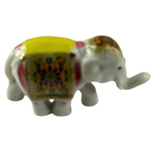 Puede incluir: Pequeña figura de elefante de cerámica blanca con montura amarilla y manta decorativa estampada. El elefante tiene detalles en rosa y un diseño colorido y detallado en la cabeza. Es un artículo decorativo en miniatura.