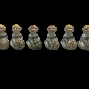Peut inclure: Six petites figurines en céramique de personnes aux cheveux jaunes et aux vêtements bleus, chacune tenant un fruit vert et brun.