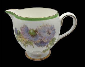 May include: White ceramic creamer with a green and gold rim. The creamer features a floral design of blue thistles with green leaves. The bottom of the creamer has a gold rim and the word "Flower" written in black.
