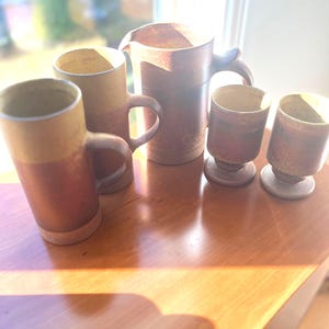 Puede incluir: Un conjunto de cerámica artesanal para beber, que incluye tazas, una jarra y tazas pequeñas. Las piezas presentan un diseño de dos tonos con una sección superior de color beige claro y una sección inferior marrón oscuro. El conjunto está dispuesto sobre una superficie de madera.