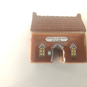 Peut inclure: Une petite maison en céramique avec un toit en briques marron et une façade marron. La maison miniature présente deux fenêtres jaunes et une porte arquée. Le texte "WHIMSEY-ON-WHY STATION" est affiché sur un panneau blanc.