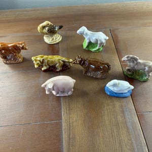 May include: A collection of small ceramic animal figurines, including a bear, duck, sheep, pig, and cow. The figurines are painted in various colors, such as brown, white, and pink. The duck is perched on a textured base. The figurines are displayed on a wooden surface.