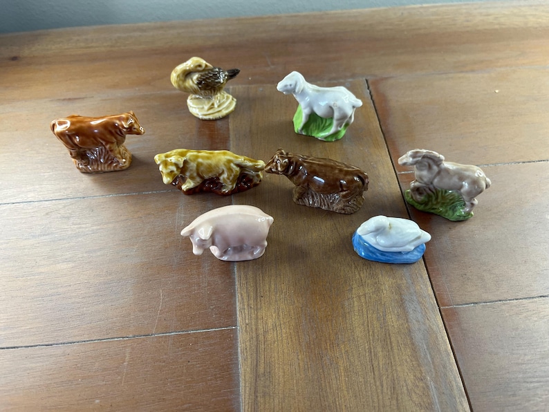 May include: A collection of small ceramic animal figurines, including a bear, duck, sheep, pig, and cow. The figurines are painted in various colors, such as brown, white, and pink. The duck is perched on a textured base. The figurines are displayed on a wooden surface.