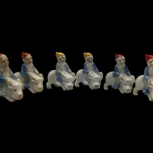 May include: Seven small ceramic figurines of gnomes riding pigs. The gnomes are wearing blue and white outfits with red, yellow, or white hats. The pigs are white with pink snouts.