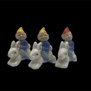 May include: Three ceramic figurines of gnomes riding white rabbits. The gnomes wear blue shirts, gray pants, and pointed hats in yellow or red. The rabbits have long ears and are in a running pose.