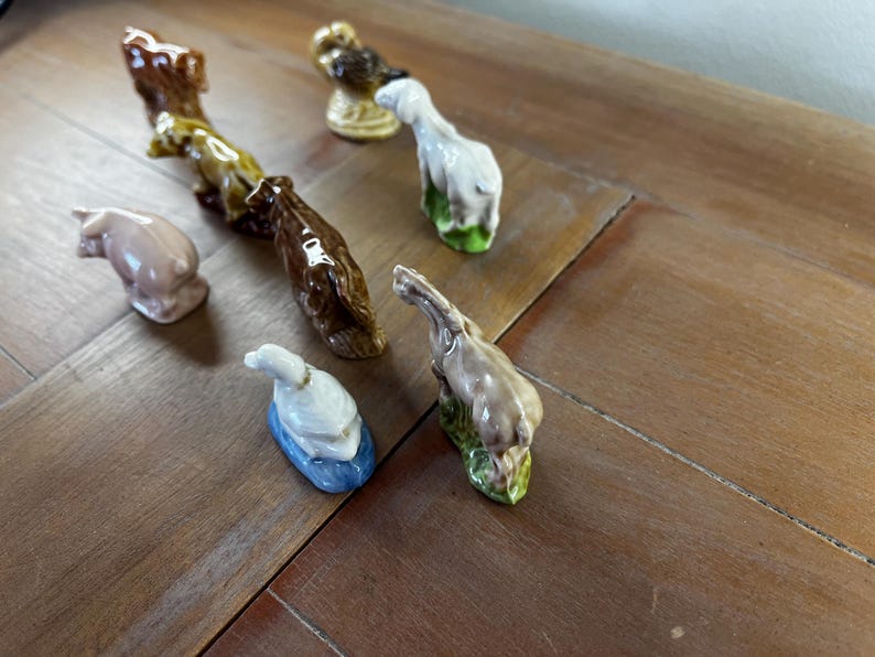 May include: A collection of small ceramic animal figurines, including a pig, a horse, a duck, and a deer. The figurines are painted in various colors, such as brown, white, and pink, and are arranged on a wooden surface.