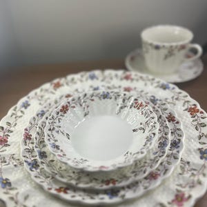 Op de afbeelding: Een witte servies set met bloemenpatroon met een kop en schotel. De set bevat een groot bord, een kleiner bord, een kom en een kopje met een bijpassende schotel. Het bloemenpatroon heeft rode, blauwe en roze bloemen met groene bladeren.
