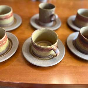 Puede incluir: Seis tazas de café y platillos de cerámica. Las tazas y los platillos son de color beige claro con una franja marrón rojizo en el medio. Las tazas tienen un asa redondeada. El juego está sobre una mesa de madera.