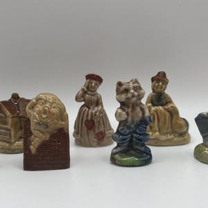 May include: A collection of eight ceramic figurines, including a woman in a red and white dress, a cat in overalls, a woman in a yellow dress, a man in a blue and brown outfit, a house, a woman lying down, and a figure with a brown and white face.