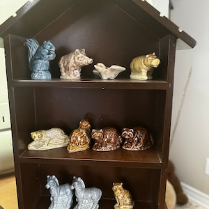 Könnte beinhalten: Ein dunkelbraunes Holzregal in Form eines Hauses mit Keramik-Tierfiguren. Die Figuren umfassen ein Eichhörnchen, ein Wildschwein, ein Nilpferd, einen Leoparden, einen Waschbären, ein Zebra und einen Tiger.