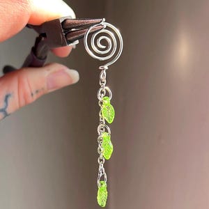 May include: A handmade silver earring with a spiral top and a chain of three green leaf charms. The earring is held by pliers against a blurred background.